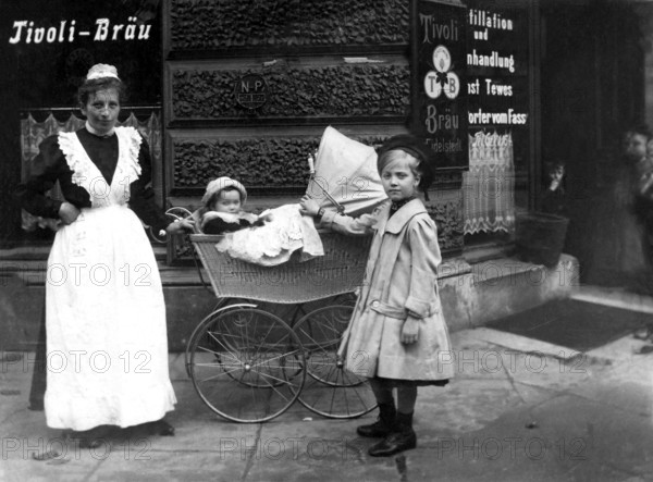 Nanny with children, ca. 1910, A woman with two children in front of a historical building with advertising signs, Historical photo