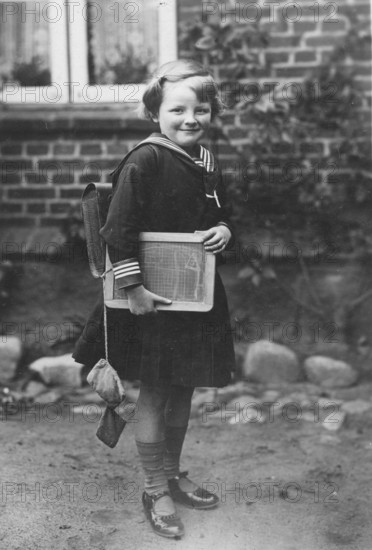 School enrolment, 1920s, A girl with a slate and school uniform stands in front of a brick wall, historical photo