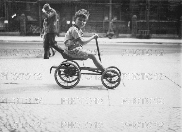 Little boy on a quadricycle, 1920s, Boy on a tricycle in an urban environment with passers-by in the background, Historical photo