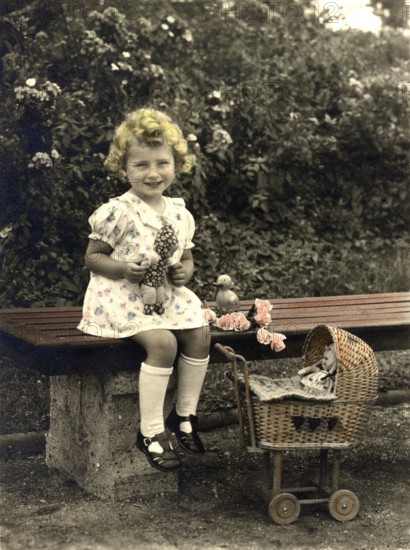 Little girl playing dolls, 1920s, Girl sitting on a bench with doll and toy trolley, surrounded by roses, Historical photo