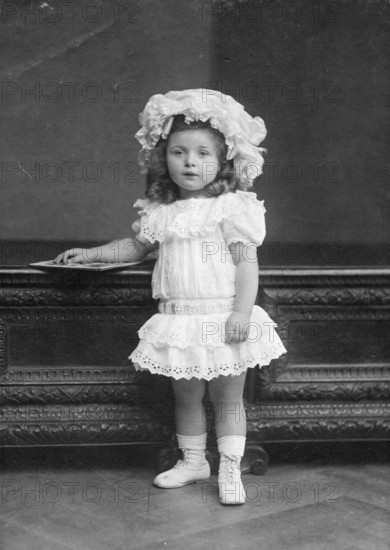 Little girl in dress, 1920s, Little girl in lace dress and hat posing in front of piece of furniture, black and white, Historical photo