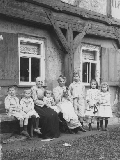 Two old ladies with children, 1920s, A large family sits and stands on a bench in front of a rustic house and poses for a photo, Historical photo