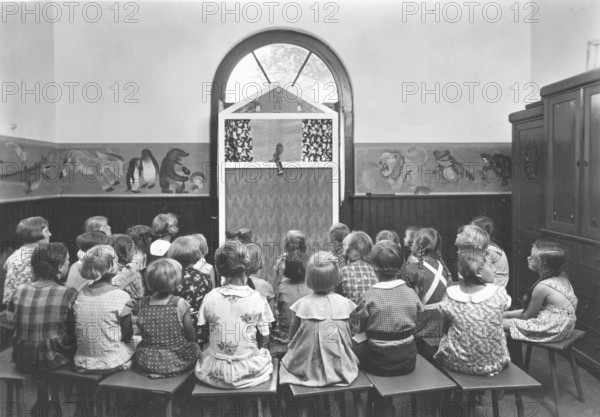 Puppet theatre, 1920s, children sit in a classroom and watch a puppet show. The walls are decorated, historical photo