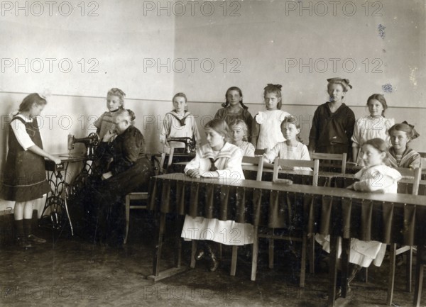 Sewing class at school, 1920, girls in a classroom practising sewing while a teacher sits at a table, historical photo