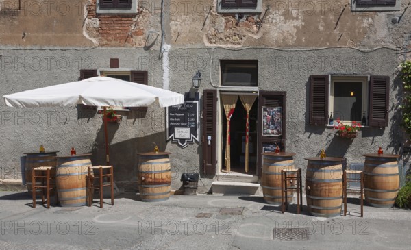 Enoteca with wooden barrels as tables and bar stools under a large parasol in front of an old house facade, Bolgheri, Tuscany, Italy