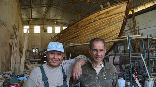 Two workers in a shipyard next to a large wooden boat hull, Diakofti boatyard, new construction of a coloured wooden boat, Diakofti, Patmos, Dodecanese, Greek Islands, Greece
