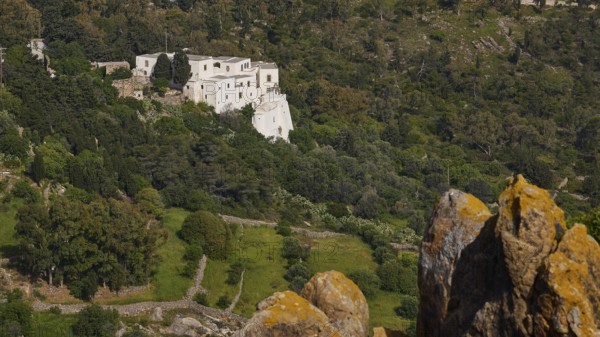 White building from a distance in a wooded landscape with rocks in the foreground, Monastery of the Revelation, Moni tis Apokalipsis, World Heritage Site 1999, Patmos, Dodecanese, Greek Islands, Greece