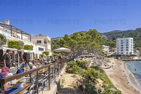 Outdoors restaurant on a terrace with tourist sitting in the sunshine and looking at the seascape view in a village by the mediterranean sea, Sant Elm, Mallorca, Spain