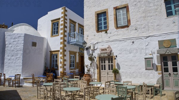 An outdoor area of a café with empty tables and chairs in front of white buildings on a sunny day, Platia Lesvias, Chora, UNESCO World Heritage Site 1999, Patmos, Dodecanese, Greek Islands, Greece