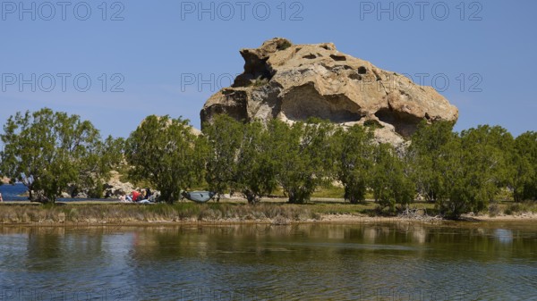 Rock rises behind a row of trees on the lakeshore under a clear sky, Rock of Kalikatsou, Seasonal Salt Lake, Patmos, Dodecanese, Greek Islands, Greece