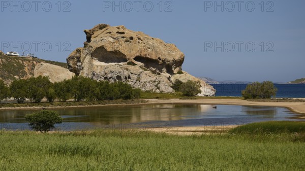 Dominant rock structure over an open water surface with green shore vegetation, Rock of Kalikatsou, Seasonal Salt Lake, Grikos, Patmos, Dodecanese, Greek Islands, Greece