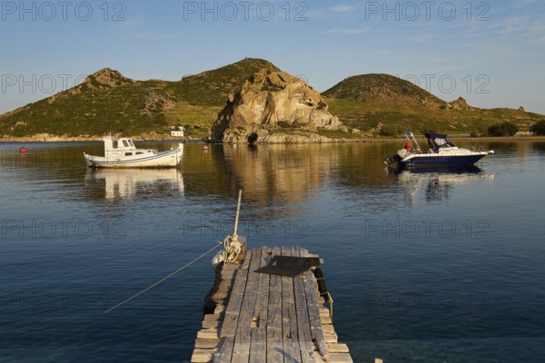 Two boats lying on calm water in front of rocky coastal landscape with wooden jetty in the foreground, Tragonisi Island, Rocks of Kalikatsou, Patmos, Dodecanese, Greek Islands, Greece