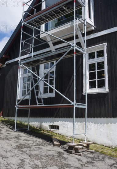 A building with white, carved mullioned windows, windows, and a black wooden facade over a white stone base on a slope, high scaffolding in front of the house, supported by wooden beams, wooden blocks and wedges, sunshine, sunny weather, work safety, construction, work, craft, danger, dangerous, unsafe, Harz Mountains, Lower Saxony, Germany