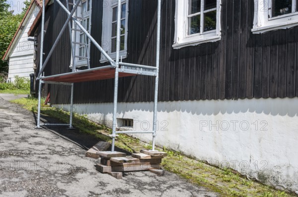 A building with white, carved mullioned windows, wooden windows, windows, and a black wooden facade over a white stone base on a slope, scaffolding in front of the house, supported by wooden beams, wooden blocks and wedges, sunshine, sunny weather, work safety, work, craft, construction, danger, dangerous, unsafe, Harz Mountains, Lower Saxony, Germany