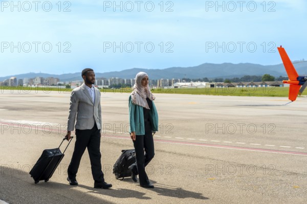 Businessman and businesswoman walking along the airfield tarmac, pulling carry on luggage while enjoying a sunny day of travel and adventure