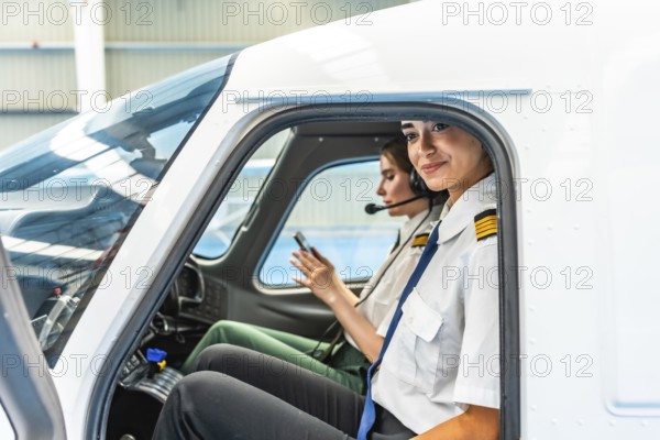 Two female pilots sitting inside the cockpit of a small airplane, preparing for take off and checking flight details on a smartphone