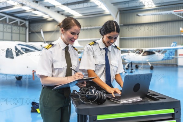 Two female pilots in uniform working on a laptop and clipboard in a spacious aircraft hangar, surrounded by small planes
