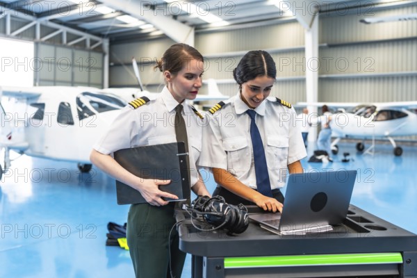Two smiling female pilots in uniform working on a laptop inside a hangar, collaborating next to an airplane, showcasing their aviation expertise
