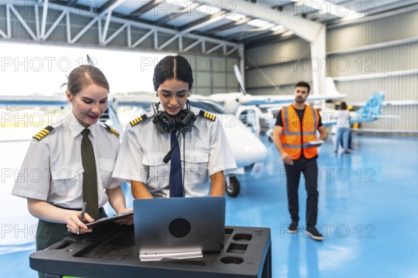 Two female colleagues pilots preparing flight plan using laptop and writing on clipboard in hangar with aircraft and airplane mechanic