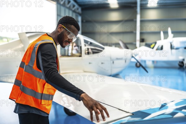 Aircraft maintenance engineer wearing safety glasses and a high visibility vest checks the wing of a small passenger plane inside an aircraft hangar