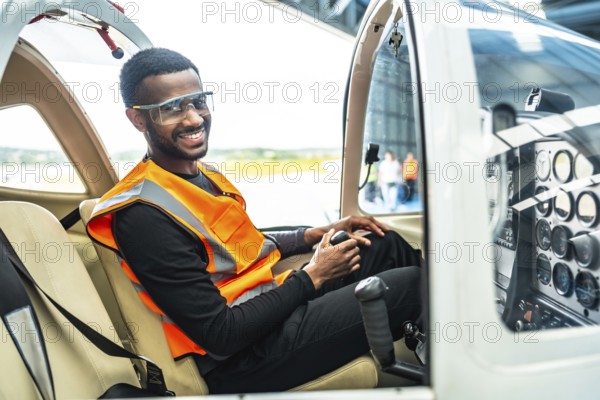 Smiling aircraft engineer sitting in cockpit holding control stick, wearing safety glasses and vest
