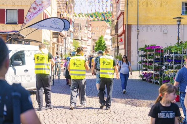 Three men in yellow waistcoats walk along a busy street, surrounded by people, Calw railway station festival, Black Forest, Germany