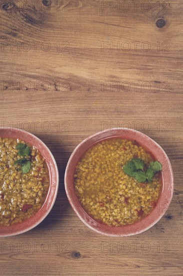 Two bowls of barley stew sit on a wooden table, each garnished with green herbs, capturing the essence of comfort food for sharing during a cozy meal