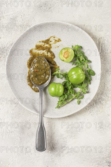A spoon with tkemali sauce, on a plate, with cherry plum and mint, top view, no people