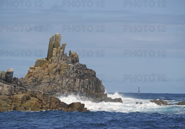 Barren rock formations by the sea with waves, a lighthouse visible in the distance, St Agnes, Isles of Scilly, Cornwall, England, Great Britain