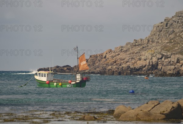 Small boat with orange sail in calm coastal waters near rocky cliff, Tresco, Isles of Scilly, Cornwall, England, United Kingdom