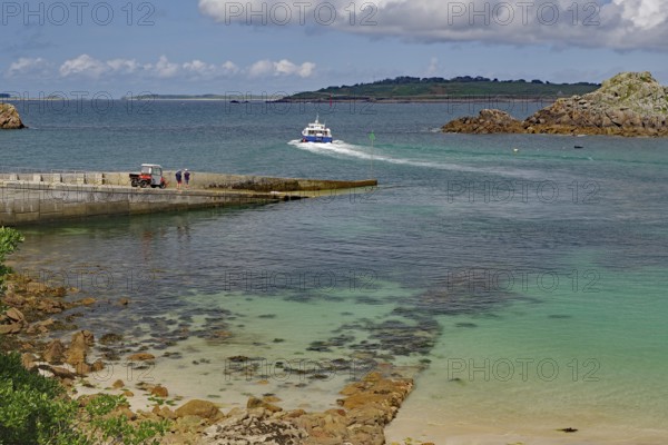 A boat approaches the harbour on clear blue water surrounded by rocks and islands, Ferry, St Agnes, Isles of Scilly, Cornwall, England, United Kingdom