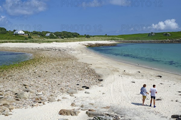 Two people walking along a soft sandy beach with blue sea and sky, St Agnes, Isles of Scilly, Cornwall, England, United Kingdom