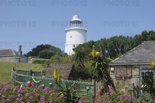 A lighthouse next to a sunny garden with blooming flowers and stone houses under a blue sky, St Agnes, Isles of Scilly, Cornwall, England, United Kingdom