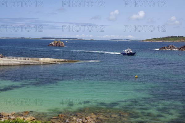 A boat sails on the open sea between small islands, under a clear blue sky, St Agnes, Isles of Scilly, Cornwall, England, United Kingdom