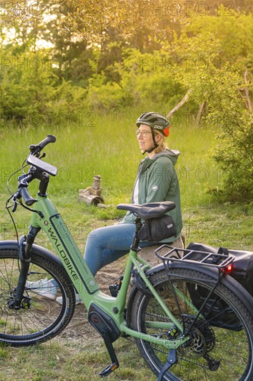Person with helmet sitting in the sunset next to a bicycle in a meadow, surrounded by nature, e-bike excursion, Gechingen, Landkreis Calw, Germany