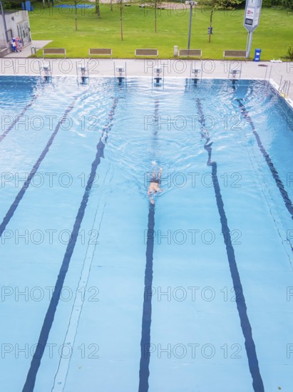 A person in a turquoise-coloured swimming pool swims laps surrounded by empty lanes on a warm summer day, Calw outdoor pool, Black Forest, Germany