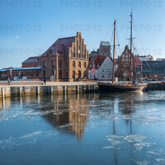Winter in the Old Harbour with historic sailing ship, ice on the water, brick architecture of Speicher, Wassertor and St. Nicholas Church, Hanseatic City of Wismar, Mecklenburg-Western Pomerania, Germany
