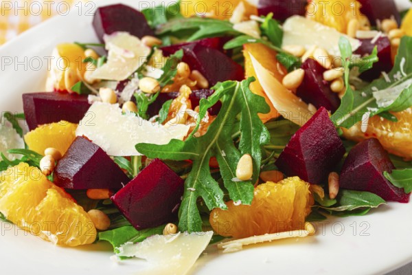 A salad made with chunks of beet, orange slices, and arugula. It is topped with cheese shavings and pine nuts, placed on a white plate with a striped napkin beside it