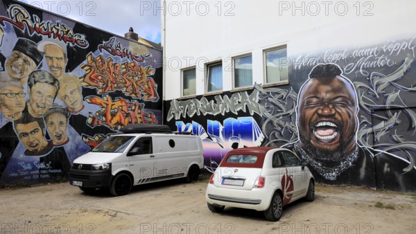 Murals of the local graffiti scene. The portrait of Keno Veith is particularly distinctive, with the saying Wenn Löppt dann Löpps, Munter. Another mural shows Dieter Bohlen together with faces that allude to the Bohlen's Right Sons theme, preceded by a Fiat 500 convertible and a VW Transport, Oldenburg, Lower Saxony, Germany