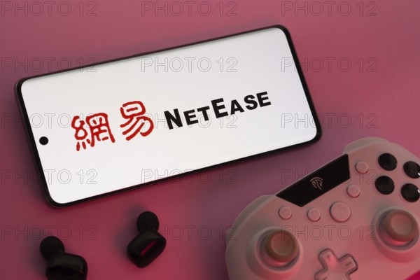 Dhaka, Bangladesh- 08 Apr 2025: A smartphone displaying the NetEase logo next to a gaming controller and earbuds on a pink surface