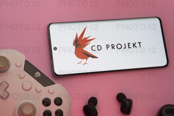 Dhaka, Bangladesh- 08 Apr 2025: A smartphone displaying the CD Projekt logo next to a gaming controller and earbuds on a pink surface