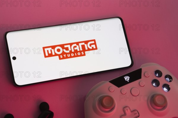Dhaka, Bangladesh- 08 Apr 2025: A smartphone displaying the Mojang Studios logo next to a gaming controller and earbuds on a pink surface