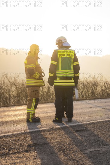 Two firefighters in protective clothing stand on a street at sunset, fire brigade exercise on the Hermann Hesse railway, Ostelsheim, Germany