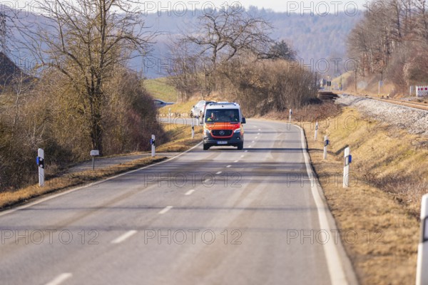An ambulance is driving on a rural road. Sparse vegetation and trees surround the road, fire department exercise on the Hermann Hesse Railway, Ostelsheim, Germany