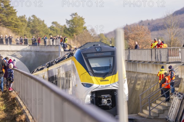 A yellow and white train travels under a bridge with spectators. Staff wearing safety vests standing on the tracks, fire department exercise on the Hermann Hesse railway, Ostelsheim, Germany