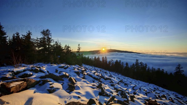 The sun sets behind a snow-covered mountain, surrounded by trees, thick fog in the valley, Fichtelgebirge