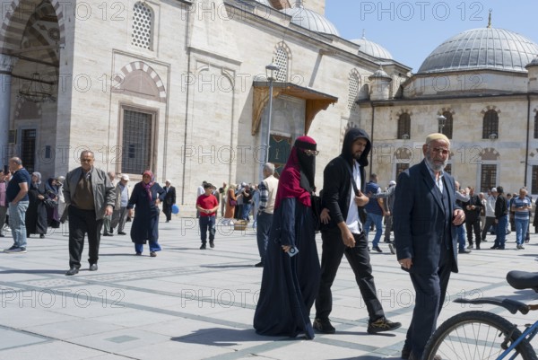 People wearing traditional clothes walking on a sunny square in front of historic buildings with domes, woman wearing burqa, Konya Sultan Selim Camii, Konya, Central Anatolia, Turkey