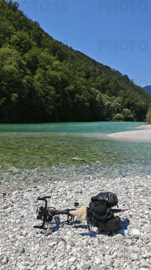 A bicycle is lying on a pebble beach by a river with clear water and wooded banks, Soca Valley, Slovenia