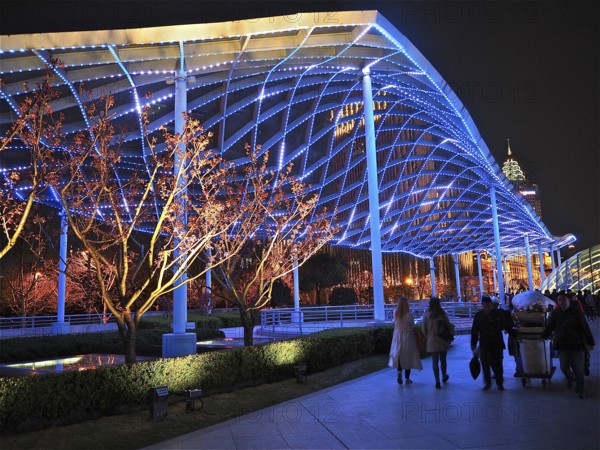 Illuminated modern structure with trees at night and people on a sidewalk, Shanghai, China