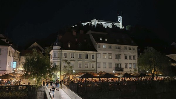 Castle illuminated at night surrounded by illuminated restaurants and visitors in an urban setting, Ljubljana, Slovenia